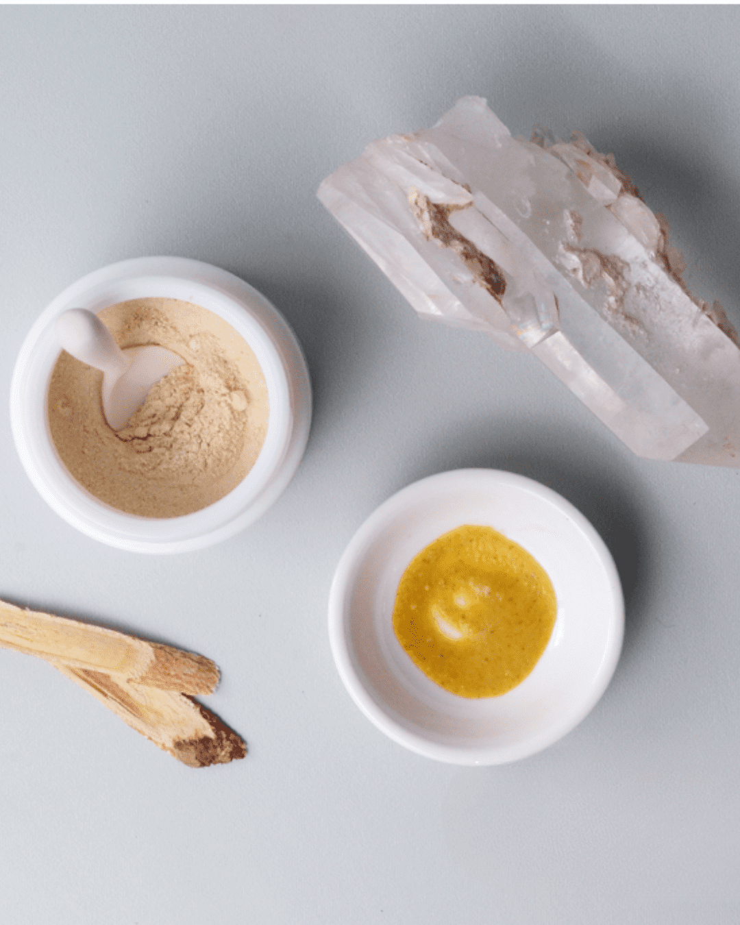 A flat-lay of holistic skincare tools, including Palo Santo sticks, a large white crystal, two ceramic bowls filled with natural powders and a turmeric-colored mixture, and a ceramic spoon arranged on a neutral surface.