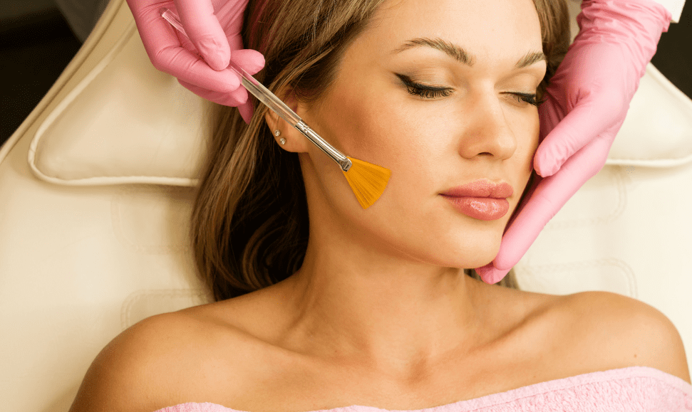 A woman with dark blonde hair and closed eyes lies on a facial table with a towel over her chest while an esthetician wearing pink gloves applies a chemical peel with a facial brush. Unlike gentler, holistic skincare treatments, this is a more intensive exfoliation method.