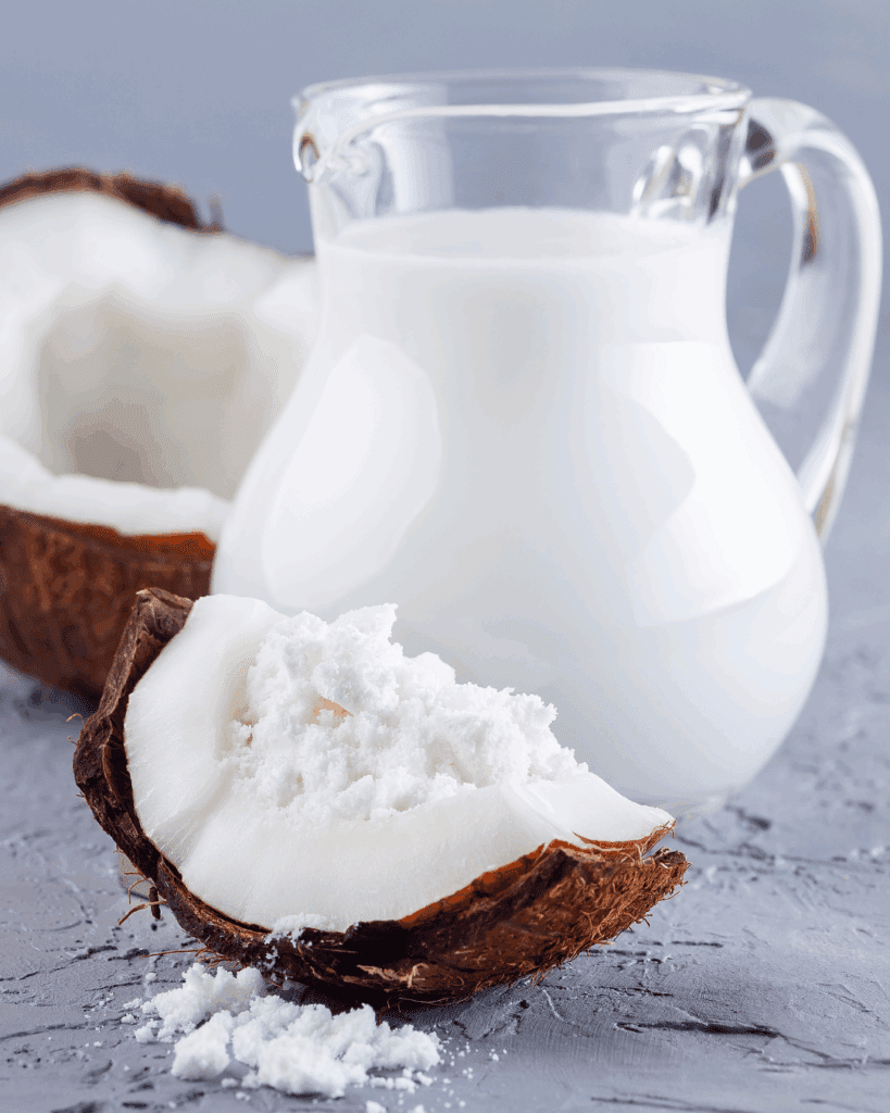 A glass pitcher of coconut water sits on a gray surface with a half coconut in the background and a quarter piece of coconut in the front, highlighting ingredients often used in holistic skincare routines.