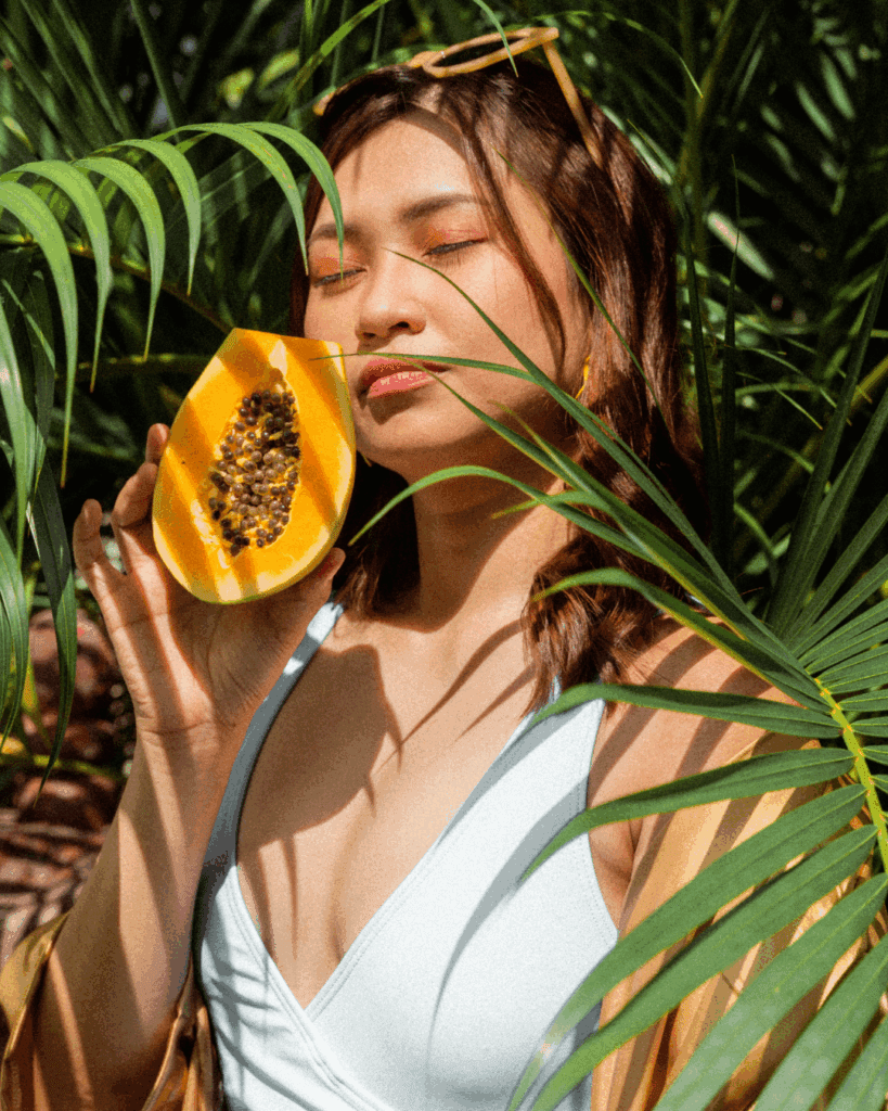 An Asian woman wearing a white V-neck tank top holds half a papaya while standing between long leafy plants outside. This ingredient is an example of a holistic ingredient you will find in Blissoma Holistic Skincare.