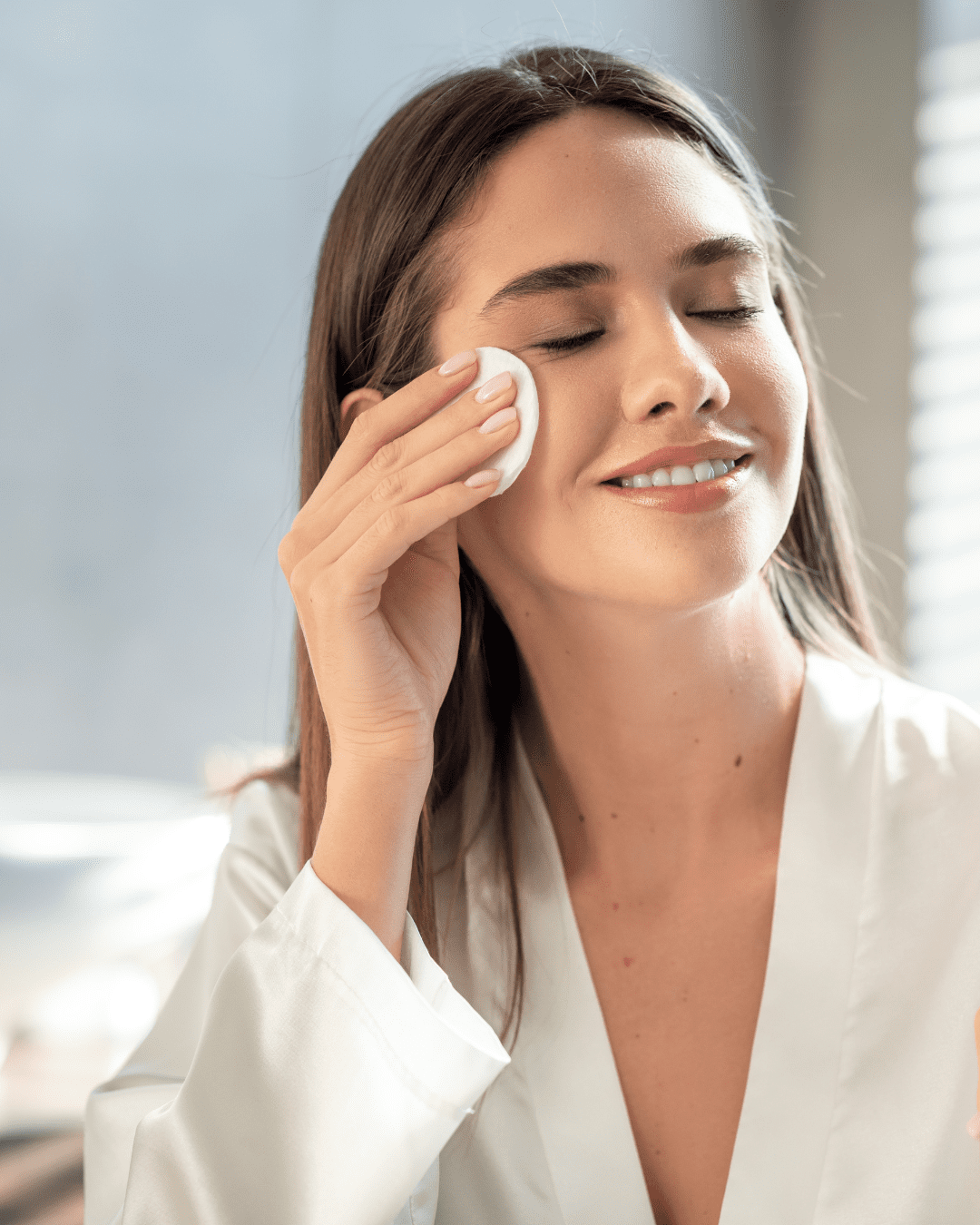 Woman wearing a white silk robe applying Algenist’s Liquid Skin Resurfacing 2% BHA Toner with a cotton round to her face, eyes closed and soft bedroom light in the background from her bedroom window.