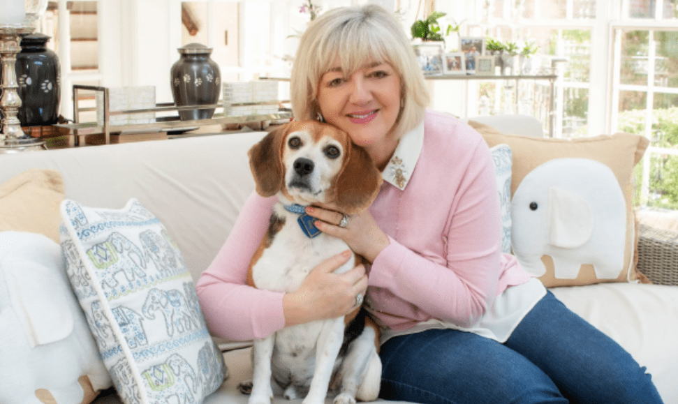 Cathy Kangas, founder of PRAI Beauty, sitting on her couch holding her beagle in her living room, with elephant-patterned pillows and memorial urns behind her, highlighting her Cathy Kangas Foundation for Animals charity.