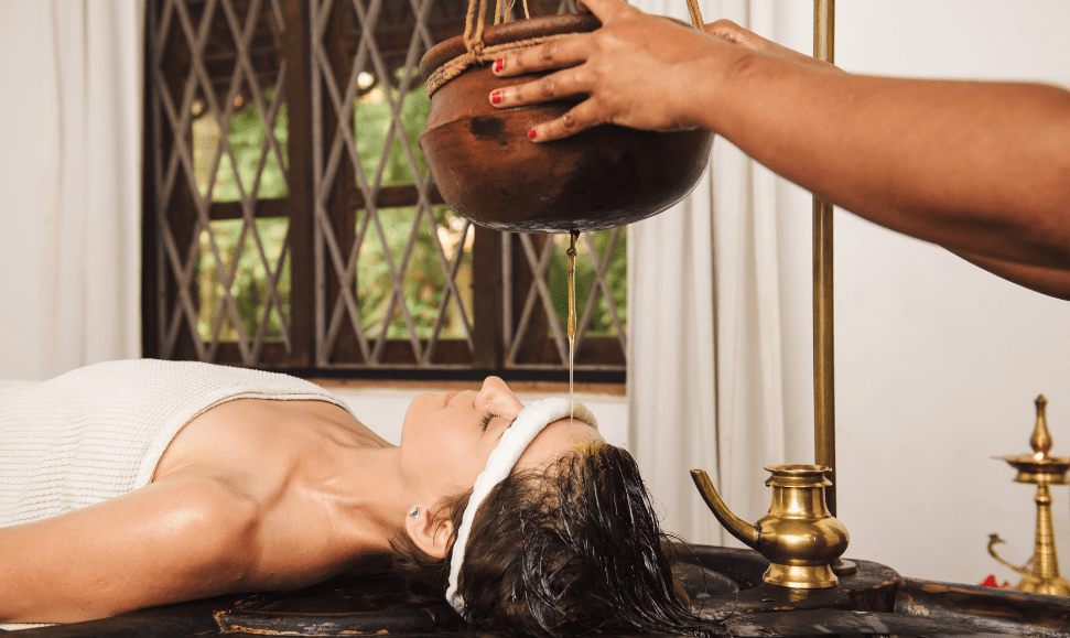 A white woman lying down with a towel wrapped around her body and a cloth headband, receiving a Ayurvedic Hair Oil Treatment. Oil drips from a hanging vessel onto her scalp while an Indian practitioner holds the vessel above her head. The spa room features a flat surface to lay on, a window with a diamond-patterned panes, and white curtains in the background.