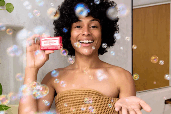 Afro-haired Black woman standing against a mustard-brown partition and white bathroom wall, wrapped in a mustard brown towel with her shoulders and upper chest exposed, holding a bottle of Dr. Bronner's Pure Castille Soap in her left hand as bubbles float in front of her.