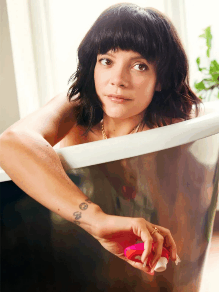 Lily Allen relaxes in a white and silver bathtub holding the Womanizer Liberty by Lily Allen, surrounded by soft lighting and a green house plant-capturing a moment of luxurious self-love and personal intimacy.