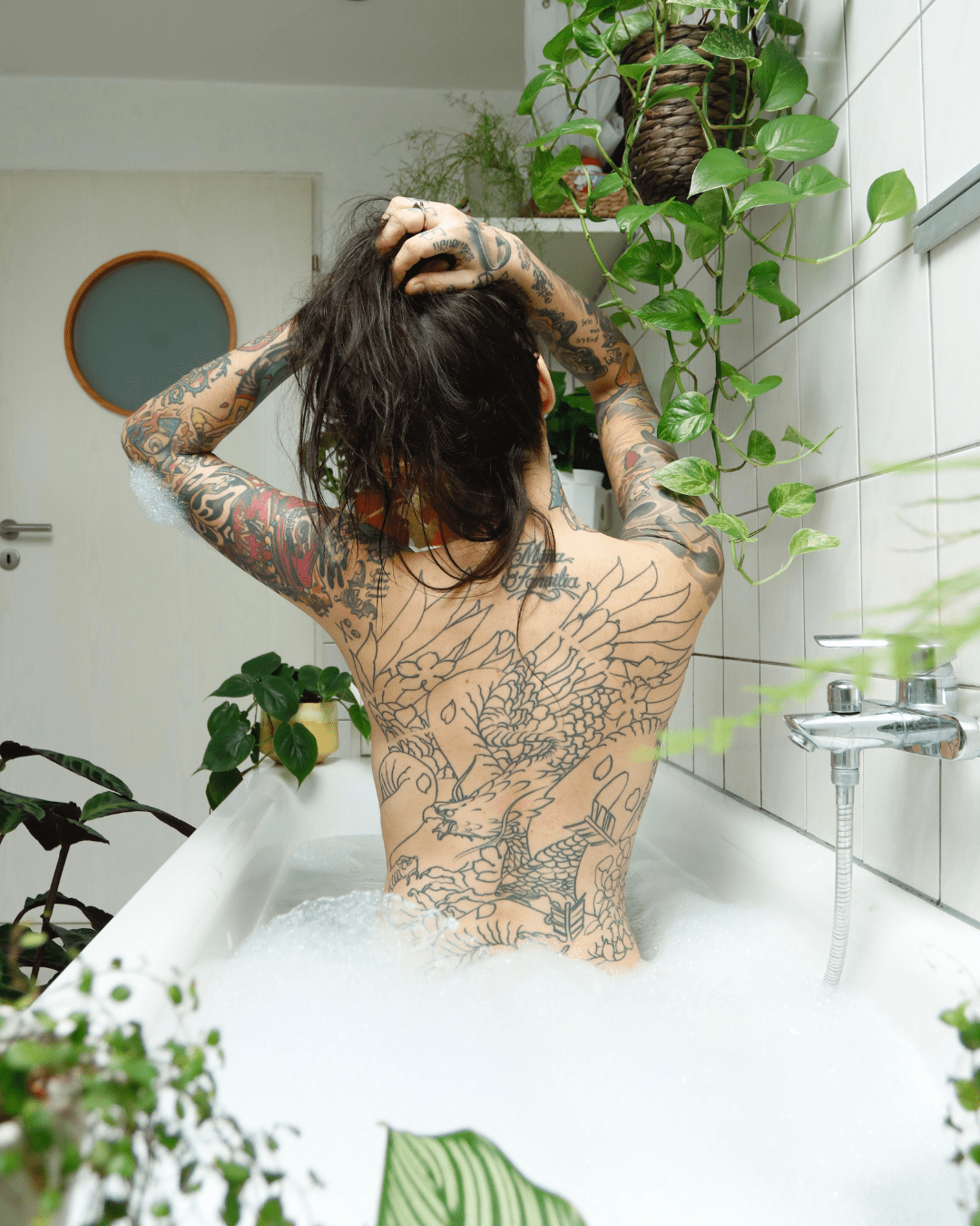 Tattoo woman relaxing in a plant-filled bathroom while soaking in a tub, capturing a sea-witch-inspired self-care moment that complements using Algenist's Liquid Skin Resurfacing 2% BHA Toner.
