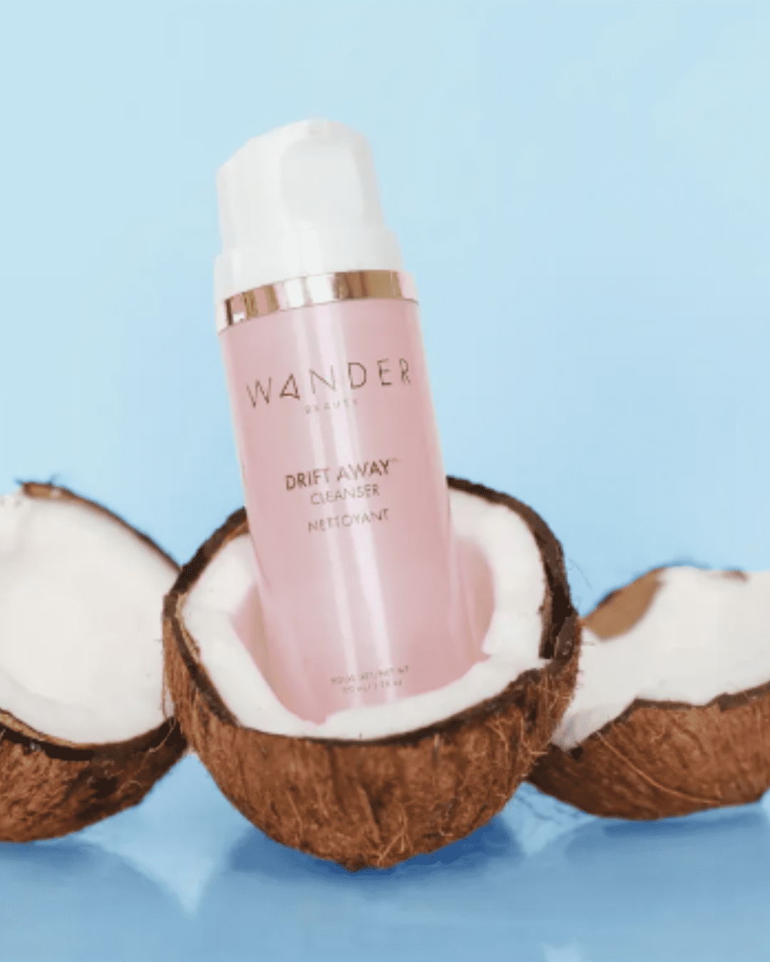 Wander Beauty Drift Away™ Cleanser in pink bottle with sliver rim and white pump, displayed with halved coconuts on reflective light blue background; a modern, safe alternative to raw egg face masks.