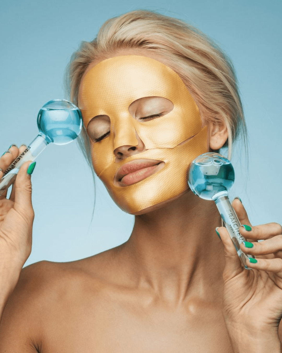 White blonde model wearing a full gold Fraîcheur Paris face mask, holding blue Fraîcheur Paris Ice Globes to her temple and jawline wiht her eyes closed, against a light blue ombre background.