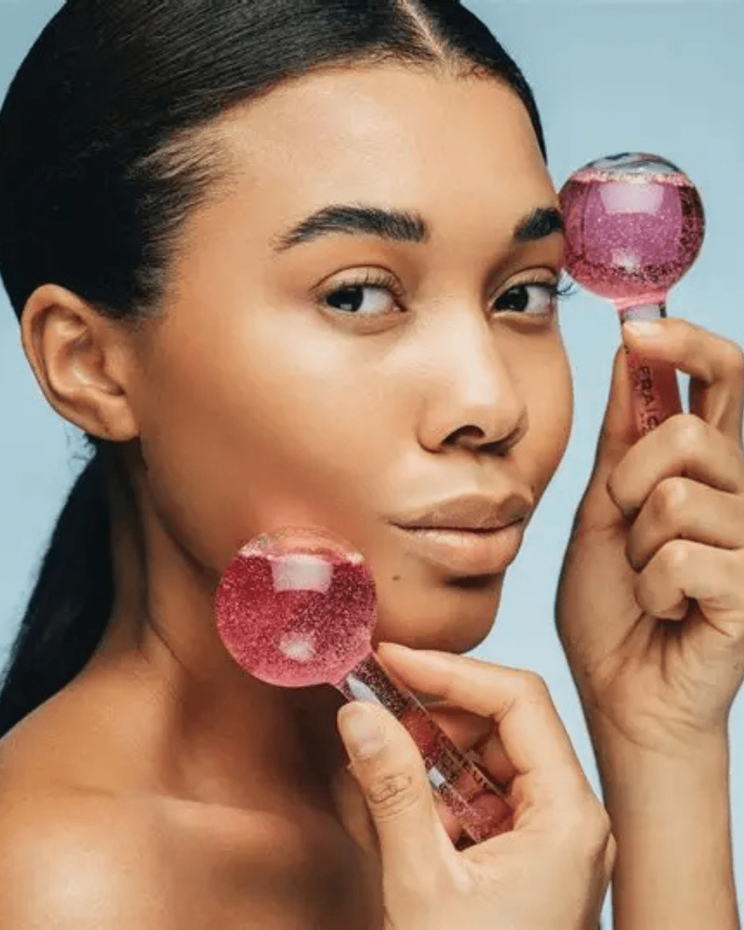 Black model with hair in ponytail holding pink Fraîcheur Paris Ice Globes, one at her jawline and one at her eyebrow, looking off camera against a light blue ombre background.