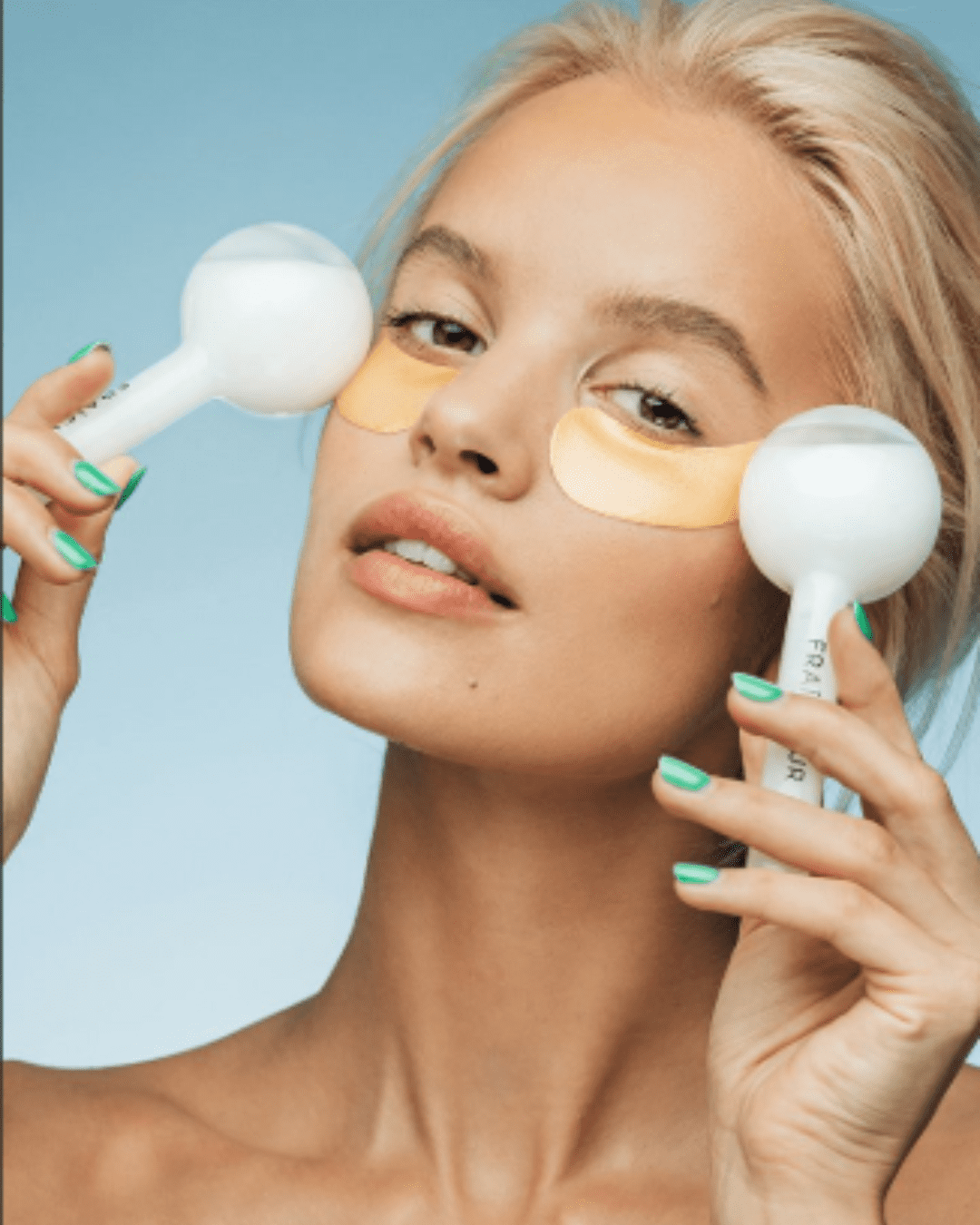 White blonde model with brown eyes and teal nails holding white Fraîcheur Paris Ice Globes to her temples while wearing gold under eye masks also from Fraîcheur Paris, against a light blue ombre background.