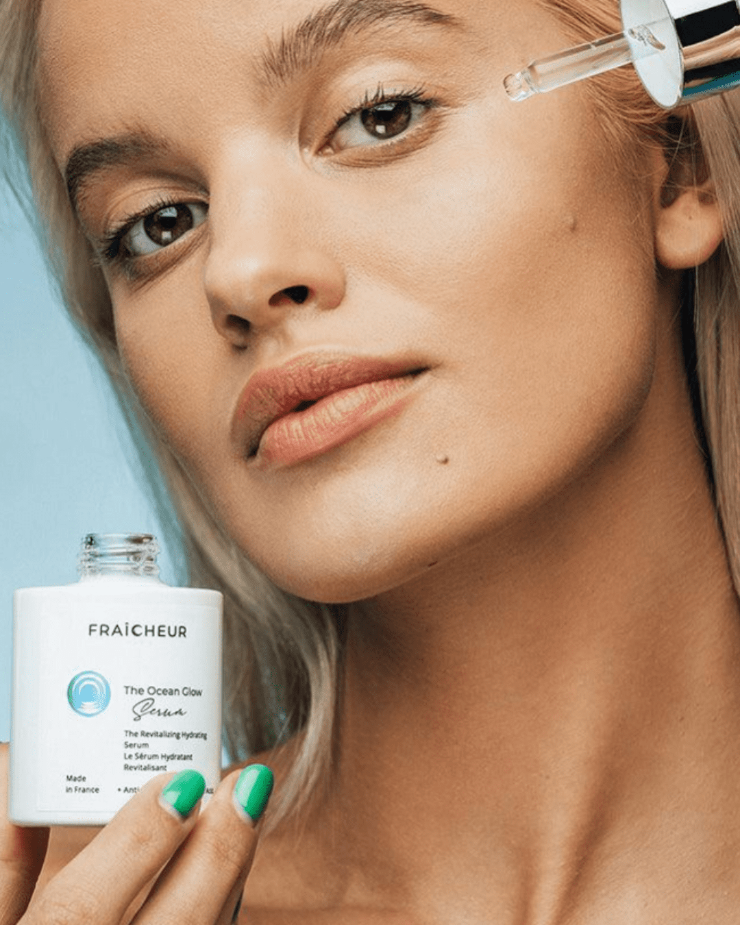 White, blonde model with green nails holding Fraîcheur Paris Ocean Glow Serum bottle and dropper near her face, a serum to be used along with Fraîcheur Paris Ice Globes, against a light blue background.