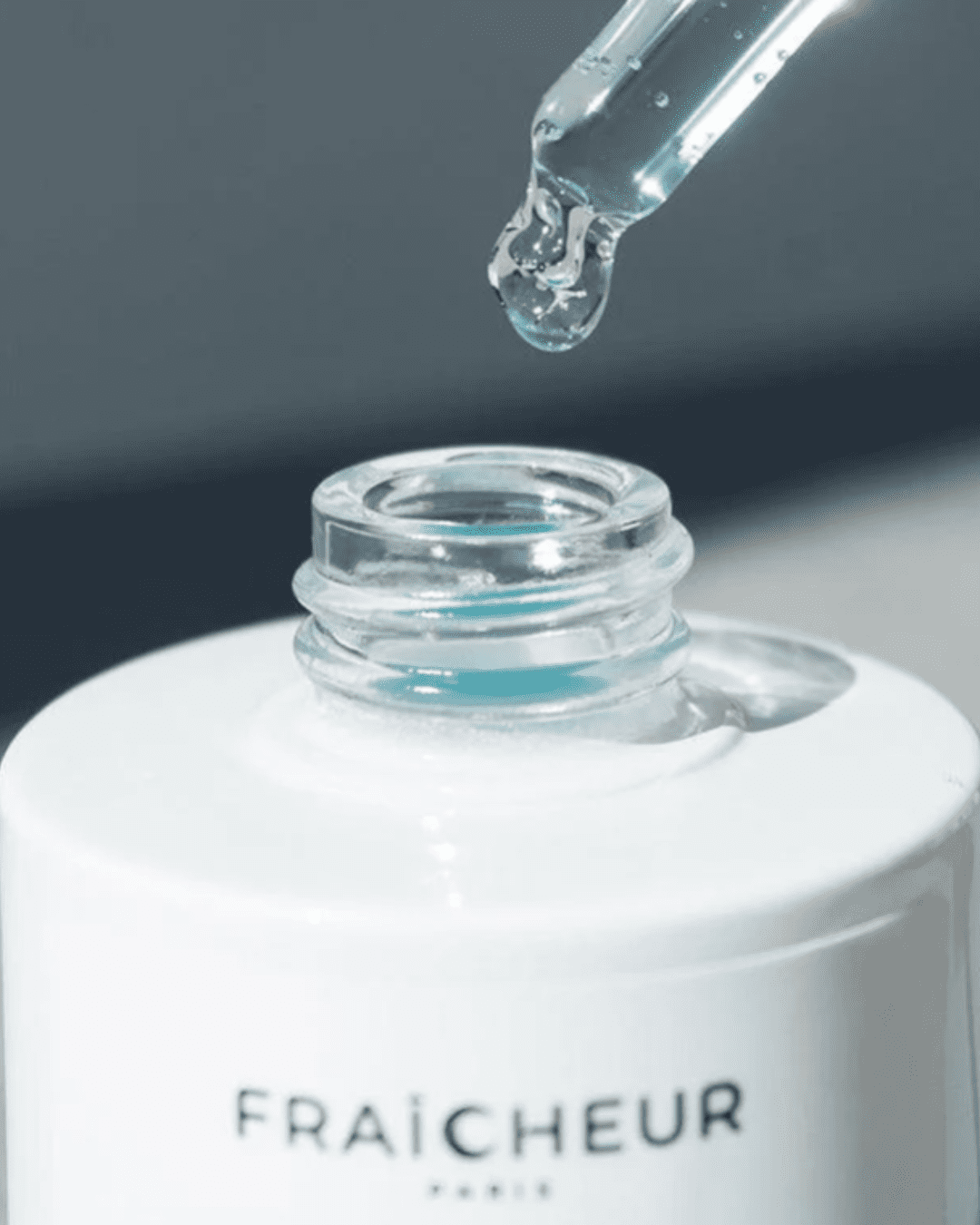 Close-up of Fraîcheur Paris Ocean Glow Serum dropper with serum dripping into the bottle, a serum to be used along with Fraîcheur Paris Ice Globes, against a soft gray, blurred background.