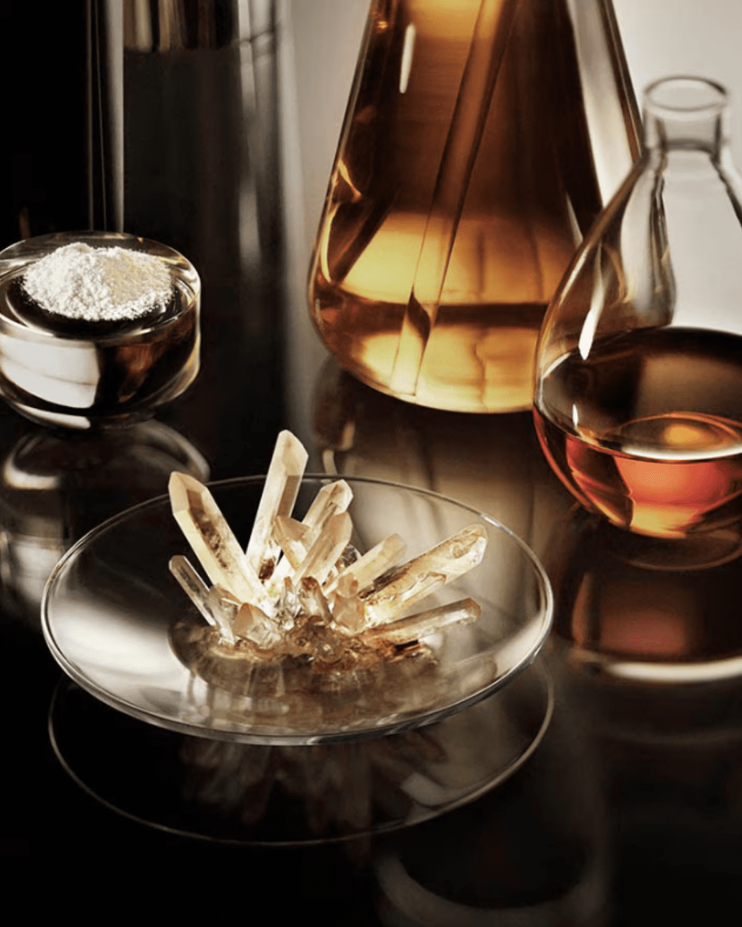 Glass beakers with liquid, a clear dish with white powder, and another dish holding a crystal, representing La Mer The Moisturizing Soft Lotion ingredients.