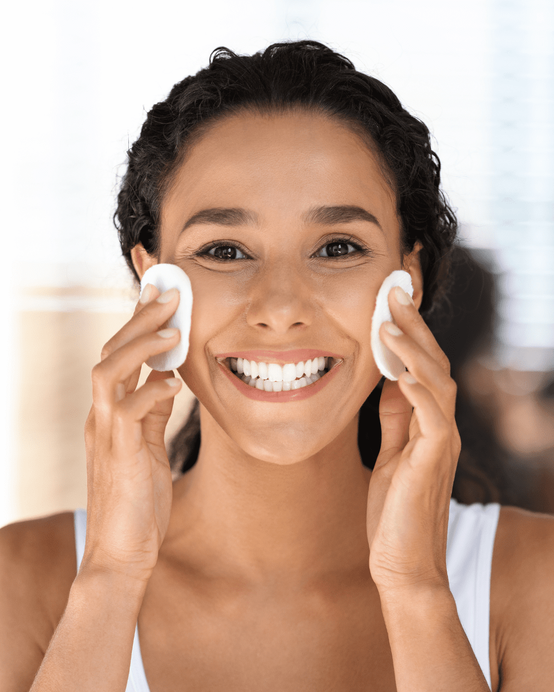 Smiling woman in white tank top playfully holding two cotton rounds to her cheeks, that could be used to apply Algenist’s Liquid Skin Resurfacing 2% BHA Toner. The background is softly blurred.