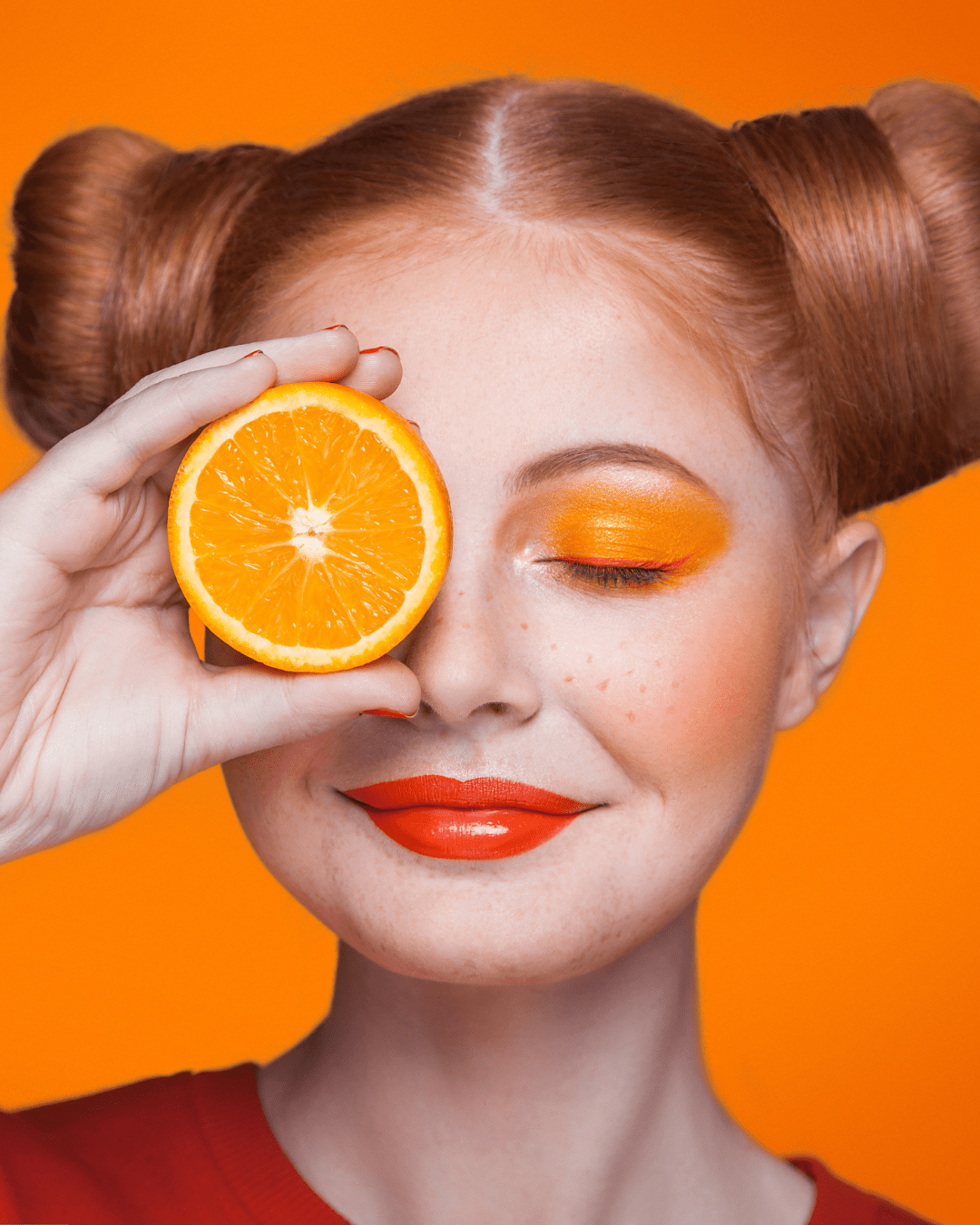 White woman with a bun on either side of her head in the style of pigtails, bright orange eyeshadow and lipstick, holding half an orange over her eye against a vivid orange background, representing Vitamin C, a key ingredient in PRAI Beauty products and in this fall self-care guide.