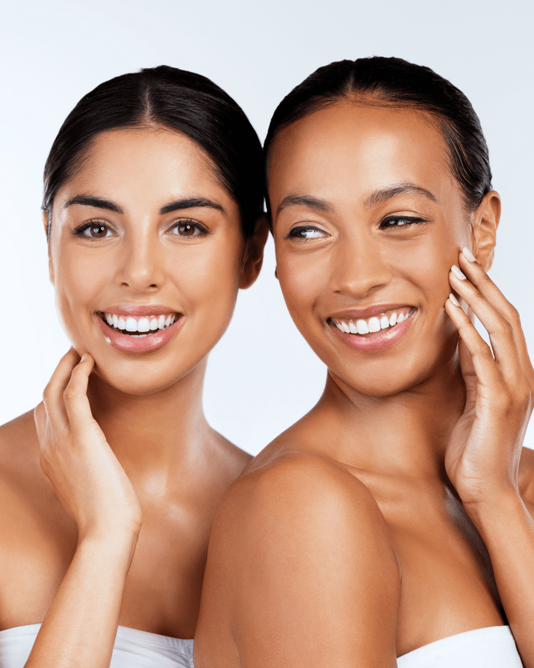 Indian woman and black girl smiling with glowing skin, a result you will get while using Algenist’s Liquid Skin Resurfacing 2% BHA Toner, on a white background, both wearing white tube tops.