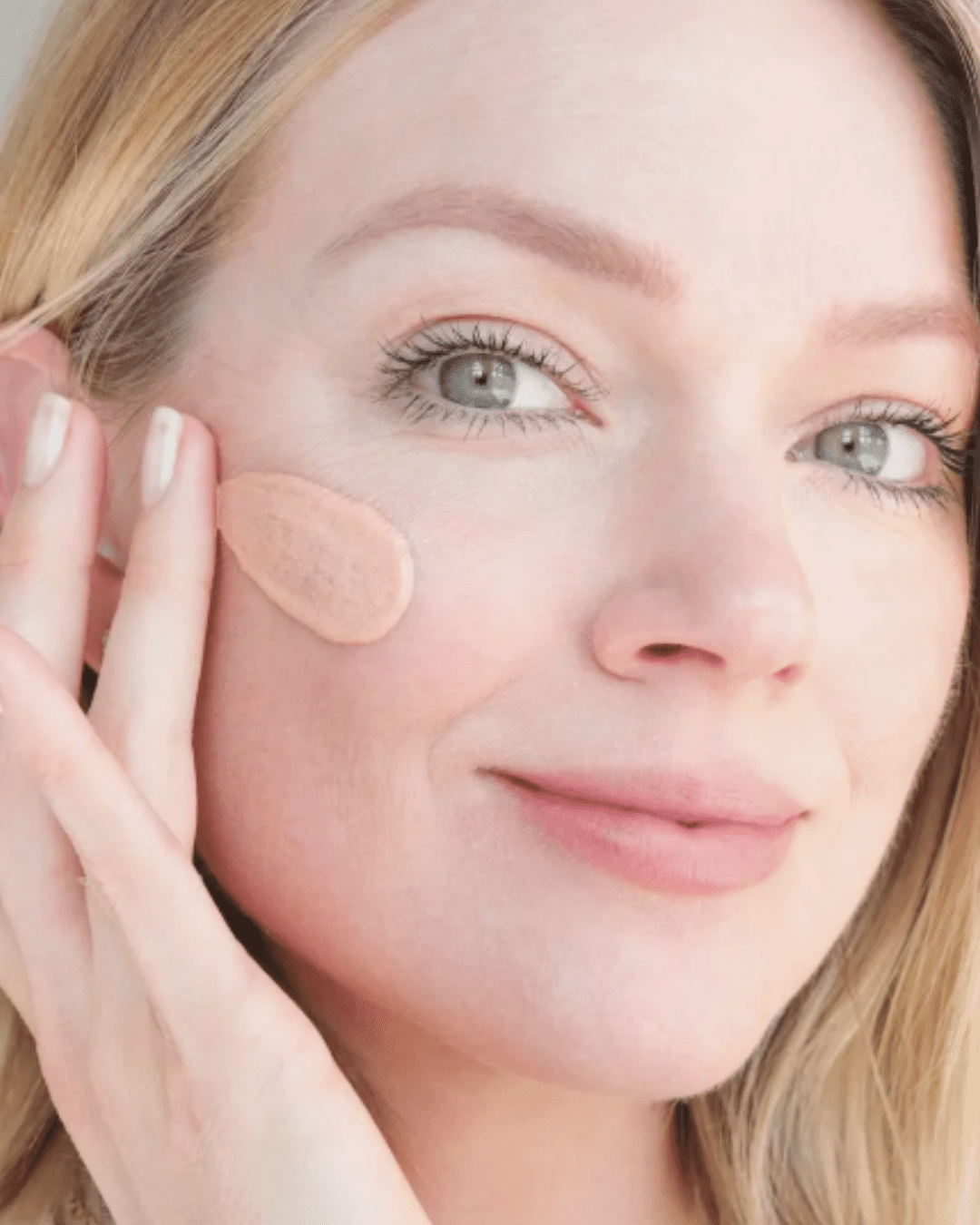 Blonde model with gray eyes smiling at camera, showing a swatch of Wander Beauty Pack Up & Glow Priming Broad Spectrum SPF 40 Mineral Sunscreen on her cheek; a modern, safe alternative to raw egg face masks.