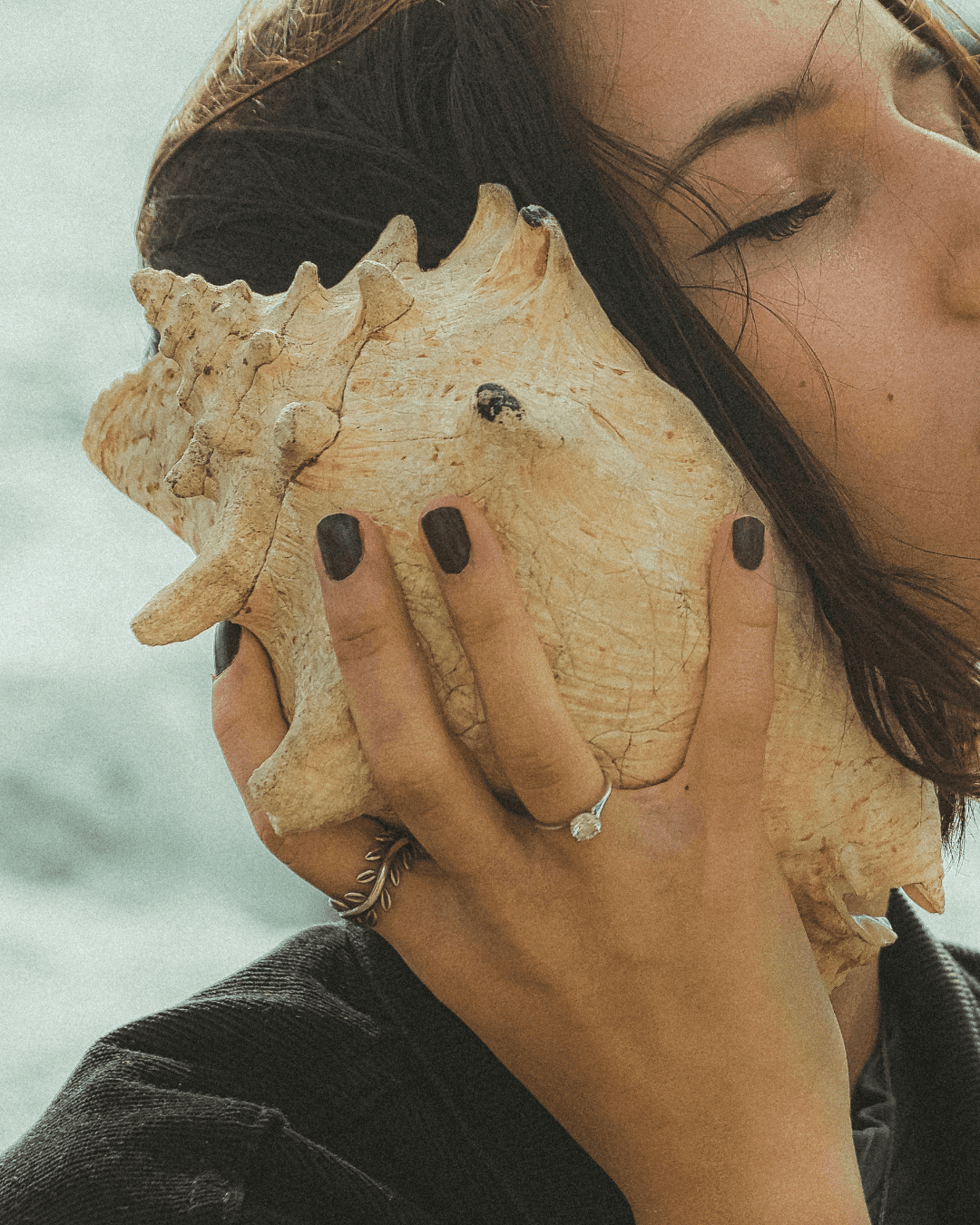 Woman with brown hair holding a seashell to her ear at the ocean, symbolizing the sea-witch ritual element of Algenist's Liquid Skin Resurfacing 2% BHA Toner routine.