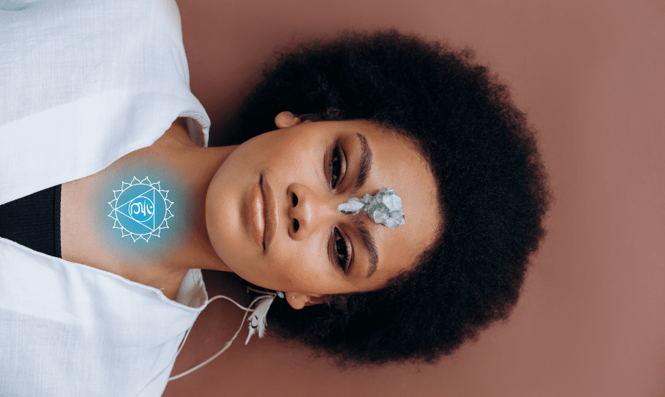Black woman with an Afro laying on a light brown surface, throat chakra symbol in white over a light blue aura on her neck, silver crystal on her forehead, wearing a white V-neck shirt over a black top.