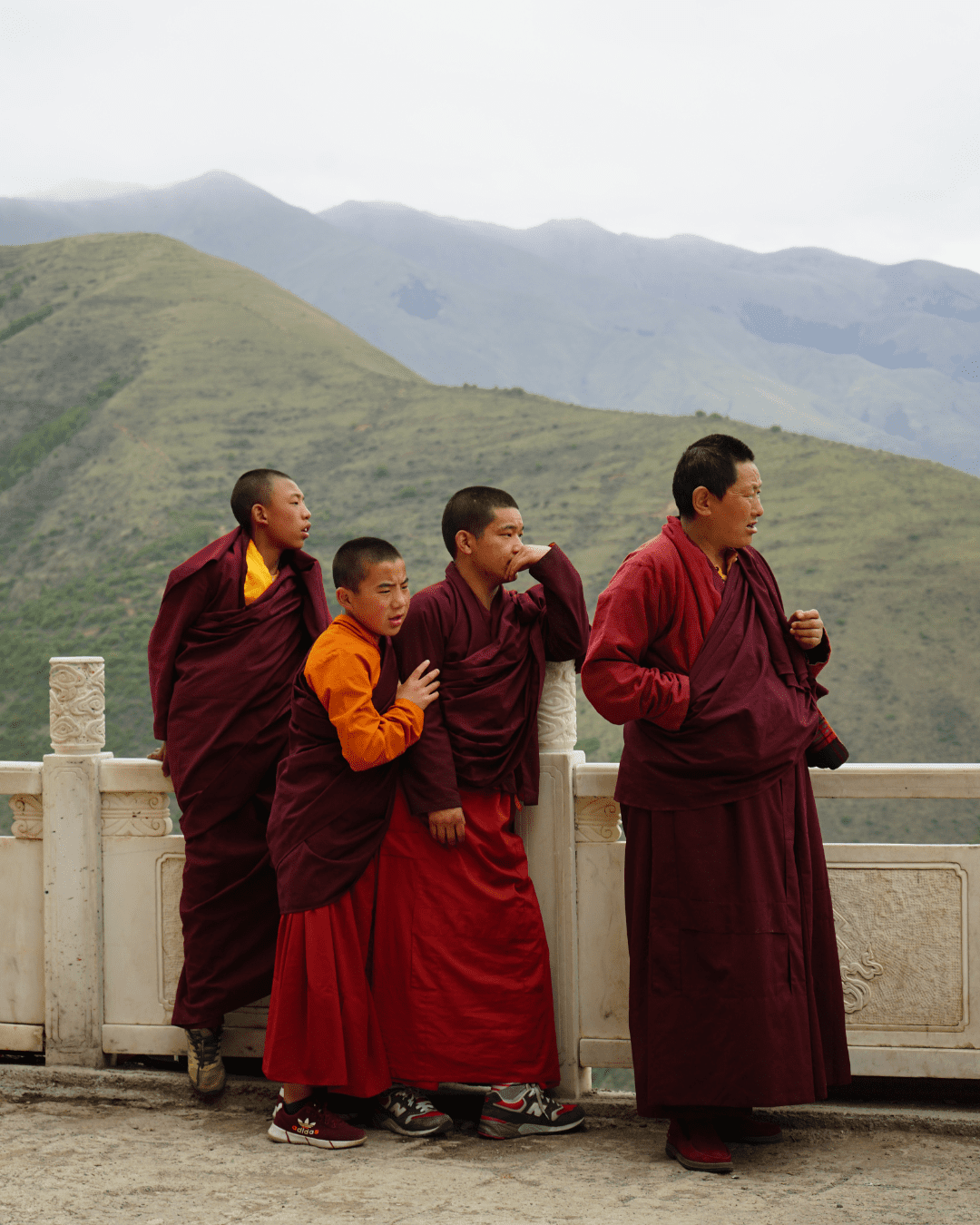 A group of Tibetan Monks gather beside a white stone fence carved with traditional markings on a dirt path. An older monk in his late 40s or early 50s, stands among younger monks who look about 8 or 9 years old. Behind them are large green hills, fading into more distant hills in the background.