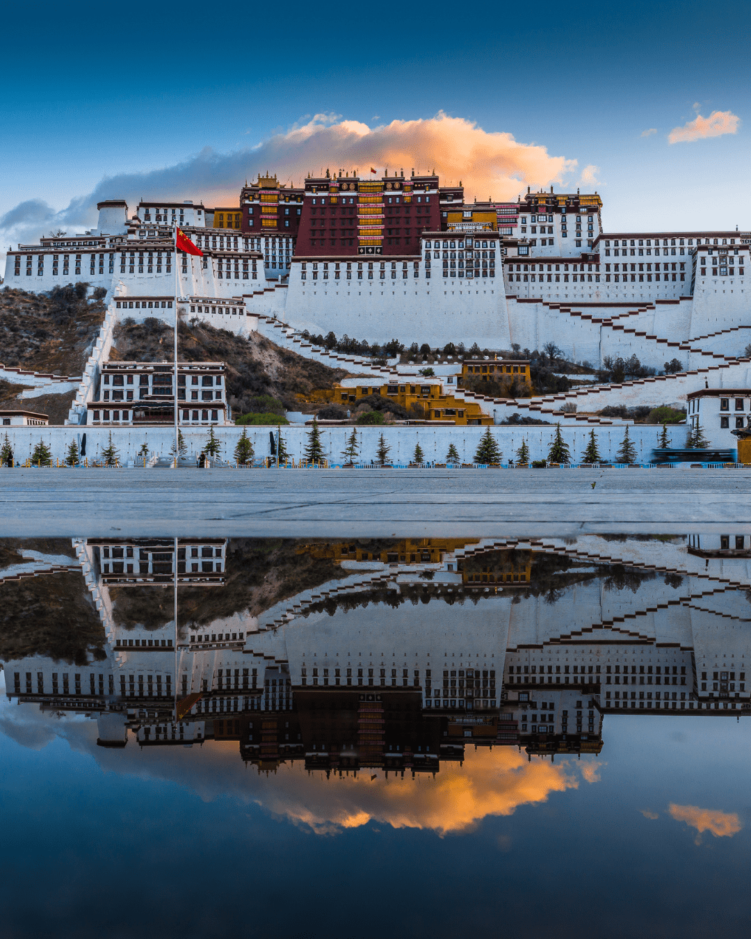 The Potala Palace in Tibet stands majestically beneath a bright blue sky scattered with soft white clouds. A calm body of water in front of the palace reflects the entire structure with mirrored-like clarity, creating a vivid double image framed by the clear, sunny day.