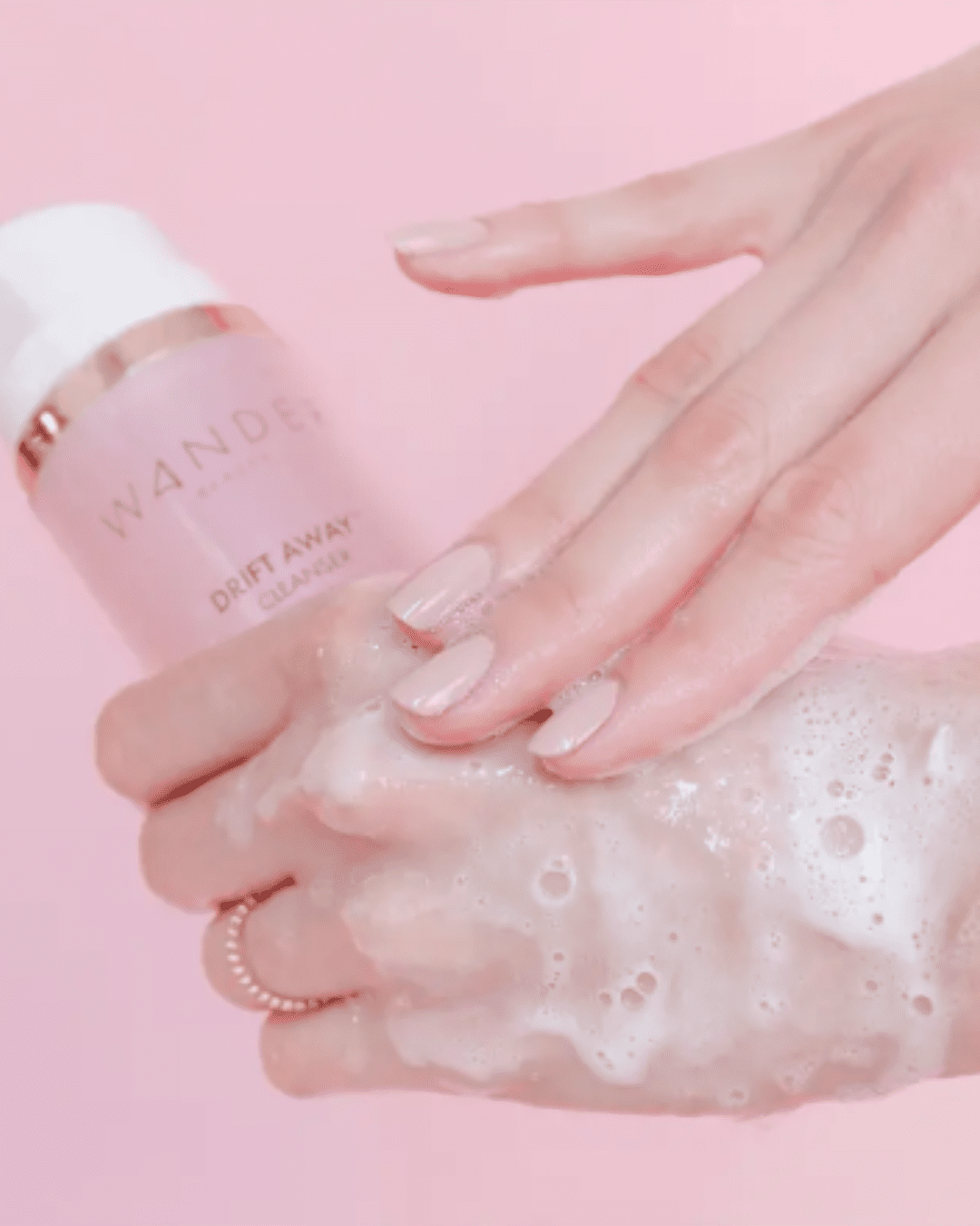 Woman holding Wander Beauty Drift Away™ Cleanser in pink bottle and silver band, with product foamed on the back of her hand; a modern, safe alternative to raw egg face masks.