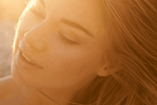 Young woman in her 20s at the beach, hair blowing in the wind, sunlight glowing behind her face, enjoying sun exposure safely.