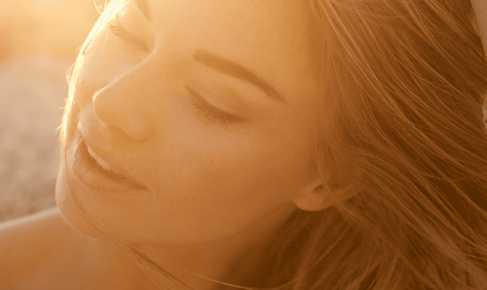 Young woman in her 20s at the beach, hair blowing in the wind, sunlight glowing behind her face, enjoying sun exposure safely.