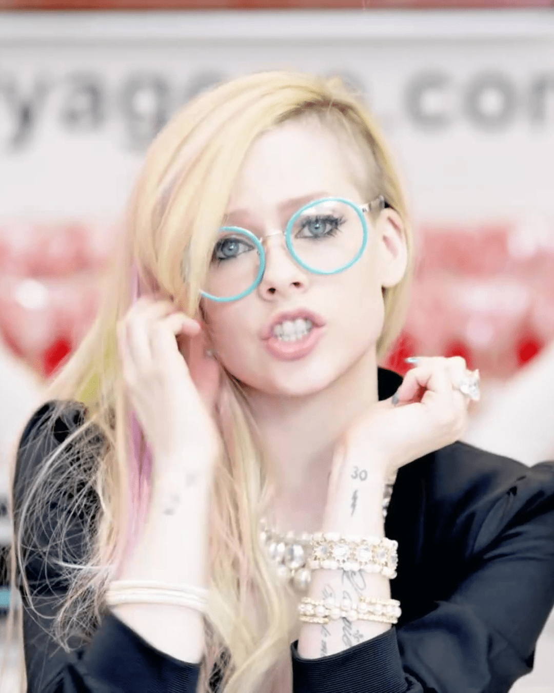 Avril Lavigne wearing light blue teal glasses and black jacket with pink streaks in her hair, wearing bracelets, still from "Hello Kitty" music video.