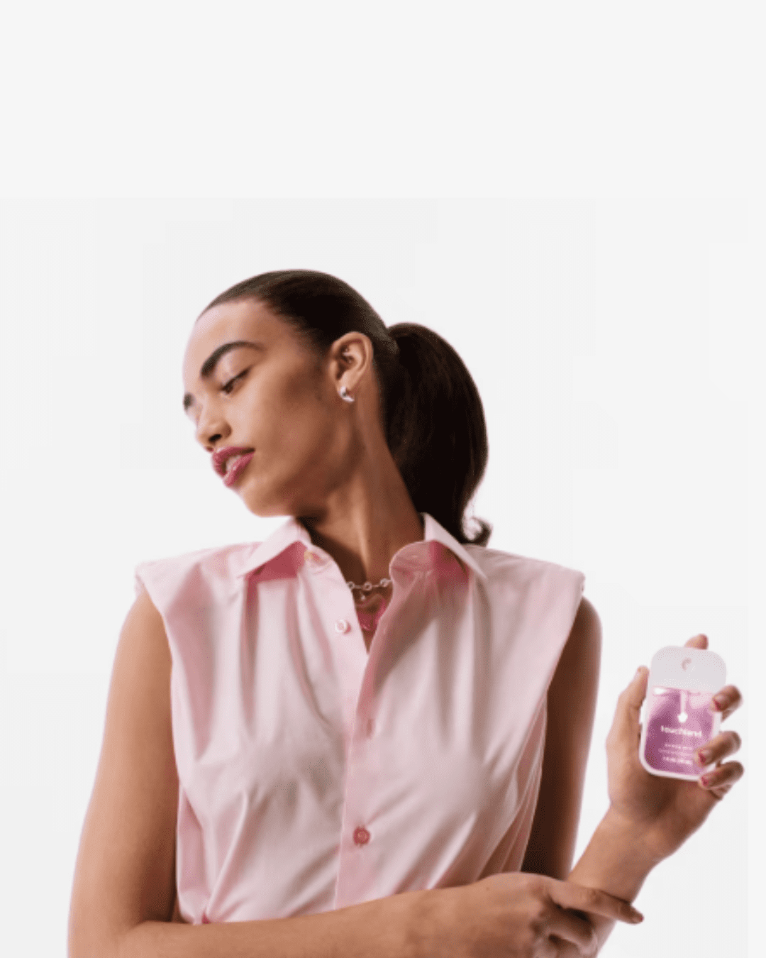 Woman with a sleek ponytail wearing pink lipstick and pink top, holding the Touchland Berry Bliss Power Mist hydrating hand sanitizer against an off-white background.