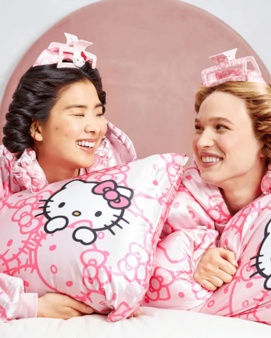 Two girls, on Asian and one White, wearing the Kitsch x Hello Kitty Satin Glazey with Hello Kitty-pattered pillows, both styled with Hello Kitty hair clips and satin curlers.
