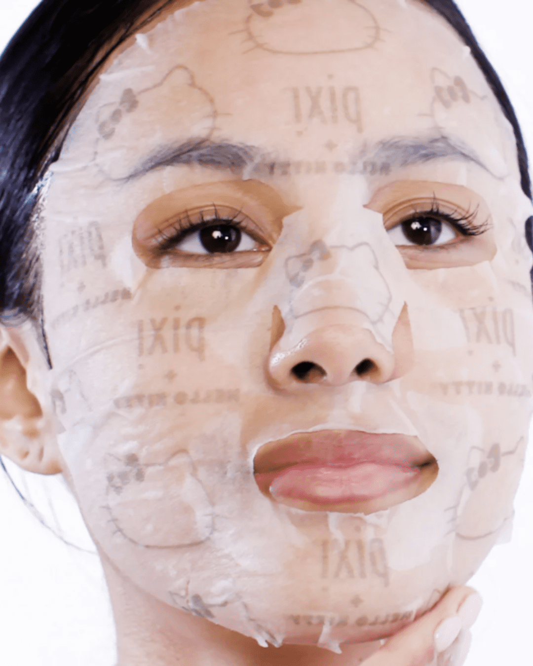 A brown-skinned woman with black hair demonstrating the Pixi + Hello "Kitty A For Apples" sheet mask applied on her face.