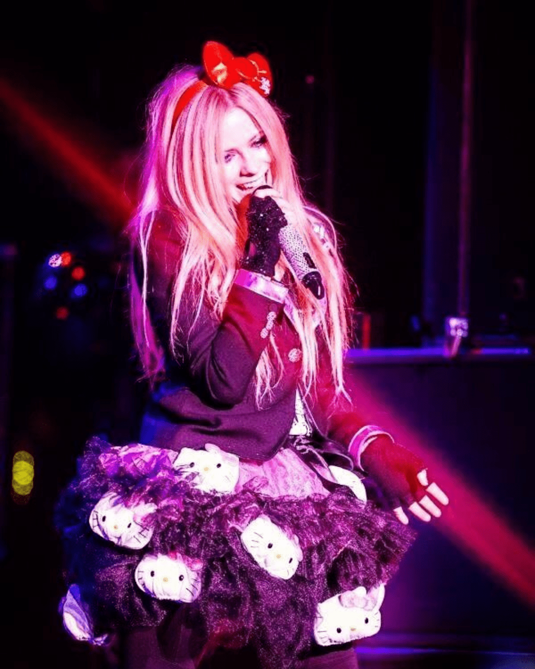 Avril Lavigne performing on stage in Hello Kitty outfit: black puffy skirt with Hello Kitty heads, red bow headband, black leather fingerless gloves, and black jacket.