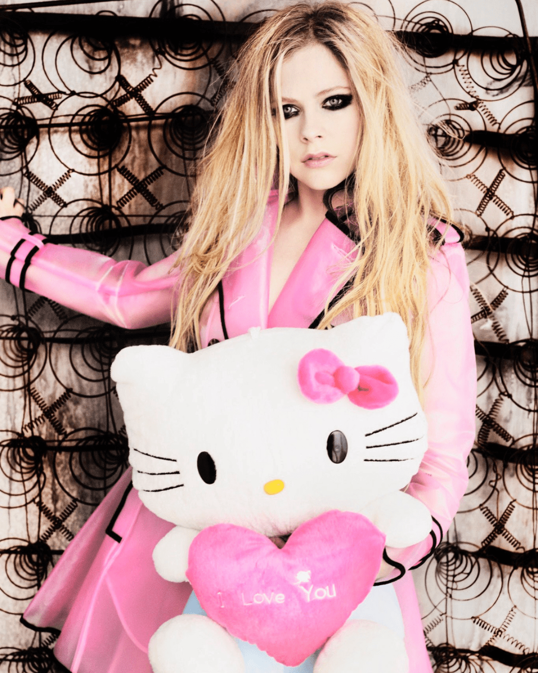 Avril Lavigne in plastic jacket dress holding Hello Kitty plush with pink heart that says "Love You", standing against background of mattress coils, heavy eyeliner.