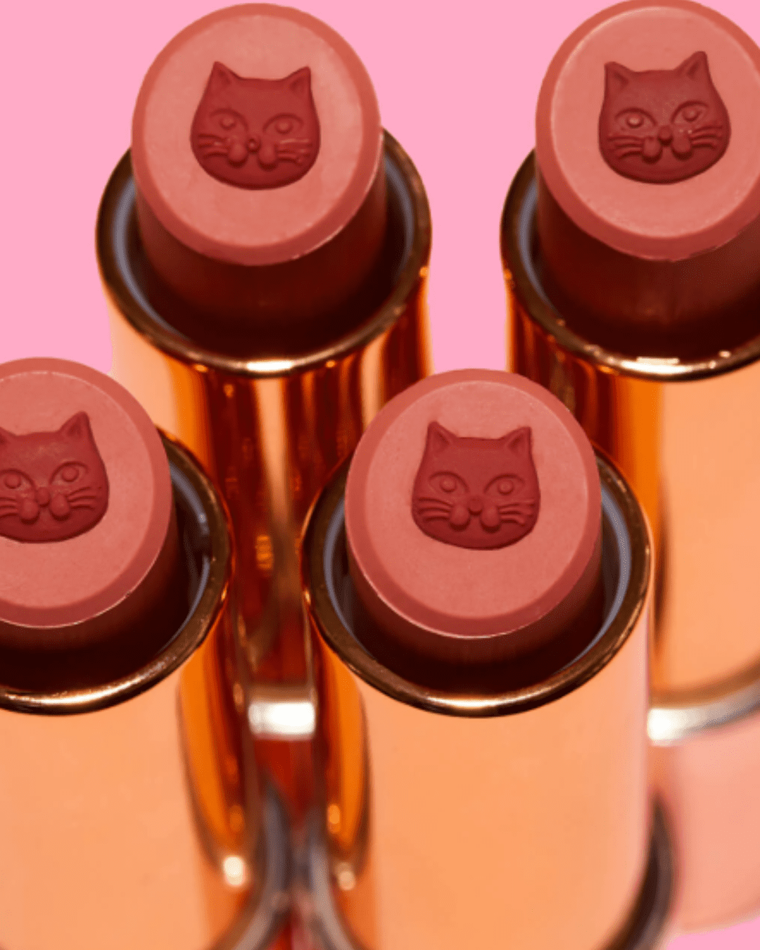 Four Winky Lux Purrfect Pout Lipsticks in the shade Pawsh arranged two on top, two on bottom, on a pink background, highlighting the gold-plated packaging and the central red kitty design in the top middle of the lipstick, capturing the playful beauty aesthetic.