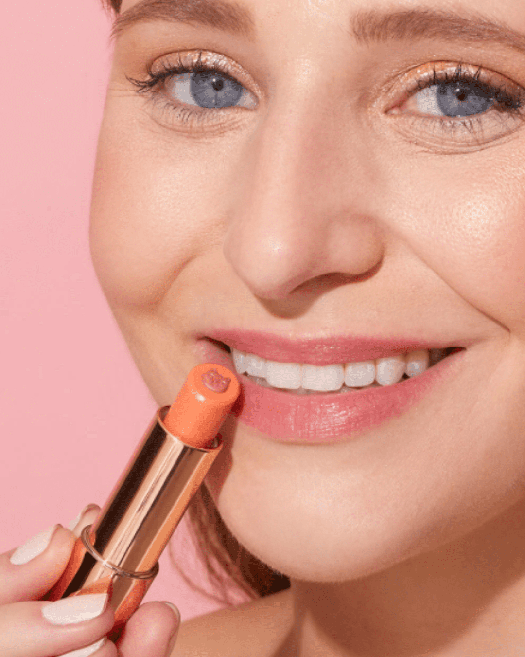 White woman with blue eyes on a pink background holding Winky Lux Purrfect Pout Lipstick shade Pounce to her lips, displaying the gold-plated beauty lipstick with the kitty design on the lipstick.