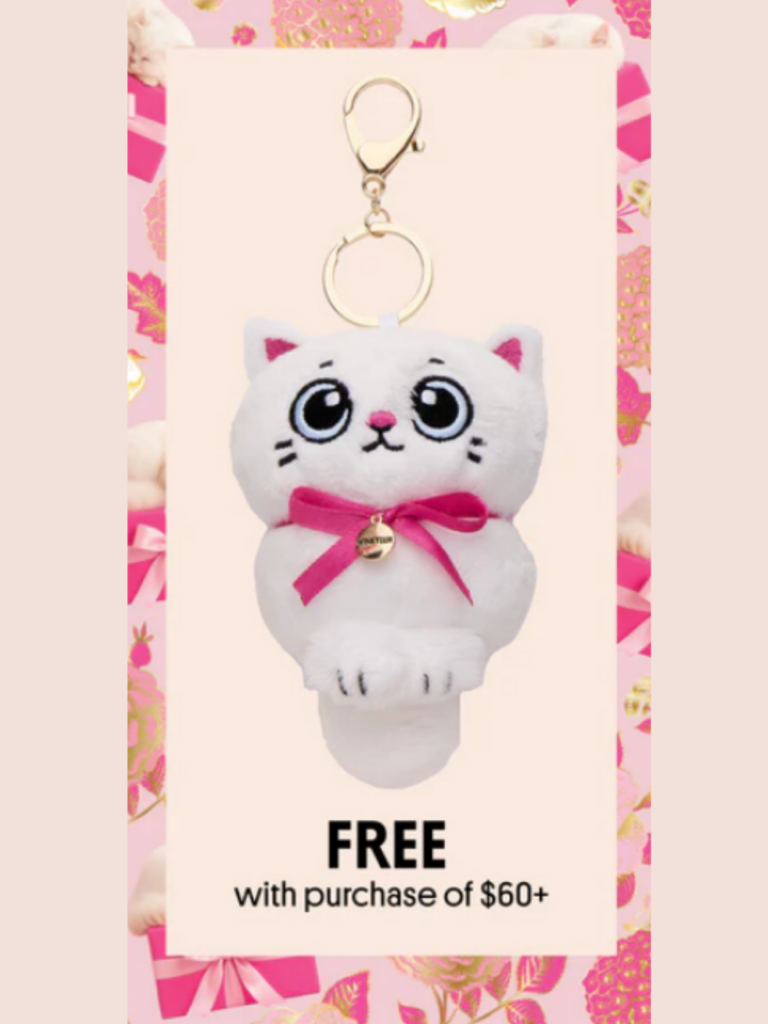 White kitty keychain with pink ears, blue eyes, and pink bow from Winky Lux, displayed against a pink floral border background, featured as a beauty gift-with-purchase for orders over $60.