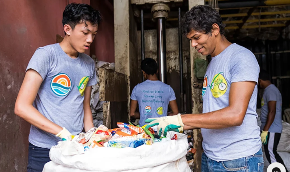 Two young men in Repurpose Global T-shirts standing on either side of a big cloth bag filled with collected plastic waste, highlighting Winky Lux's beauty sustainability partnership.