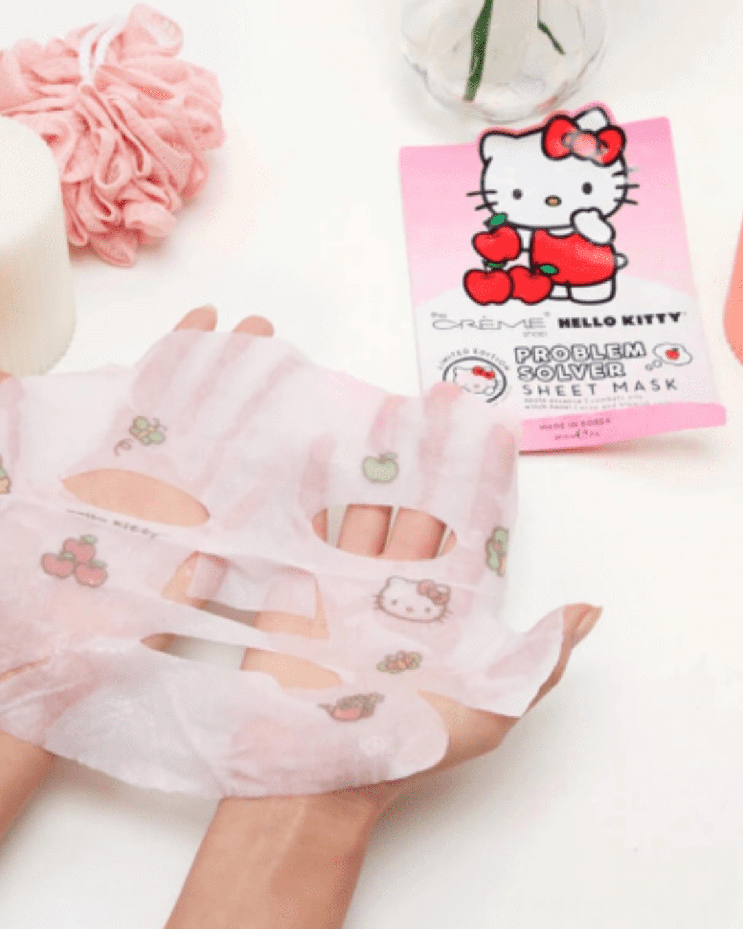 Two hands holding the Hello Kitty Problem Solver Sheet Mask fully spread out, featuring butterflies, apples, and Hello Kitty prints; packaging is in the upper right, with a glass vase, white candle and pink loofah in the upper left corner.