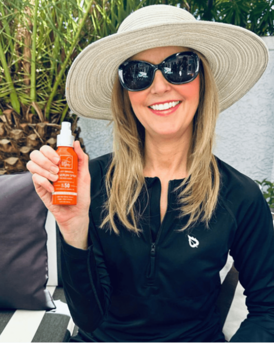 Blonde woman wearing sunglasses and a hat holding Suntegrity Milky Mineral Sun Serum Spray, Broad Spectrum SPF 50 while sitting by the pool.
