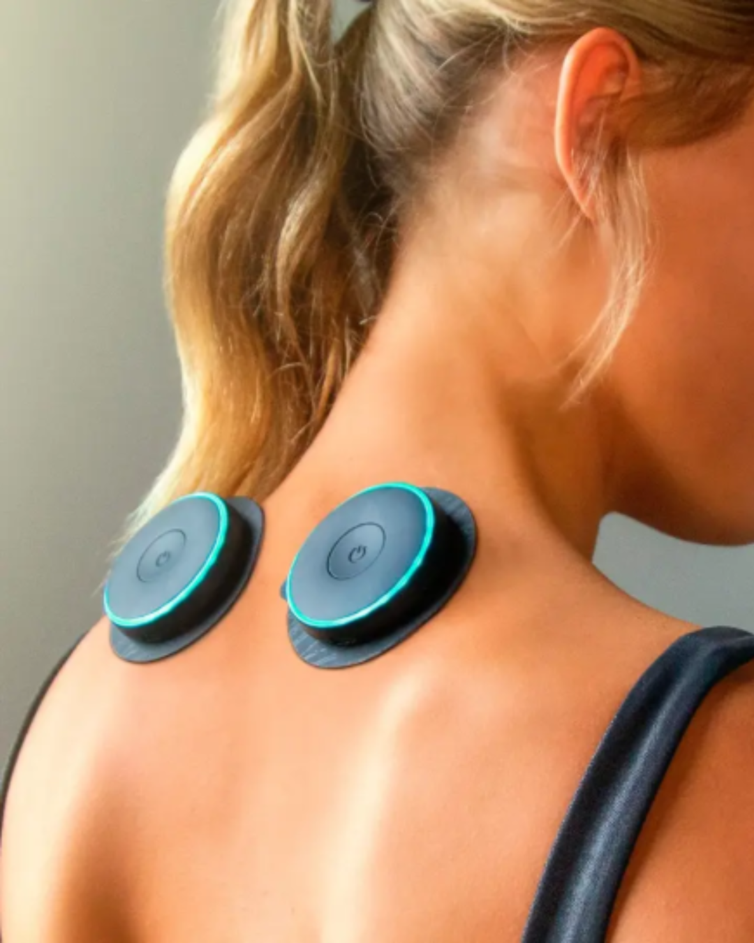 Blonde woman in a tank top using the Chirp Halo Double muscle stimulator on her upper shoulders just below her neck, demonstrating at-home muscle recovery as part of a self-love practice.
