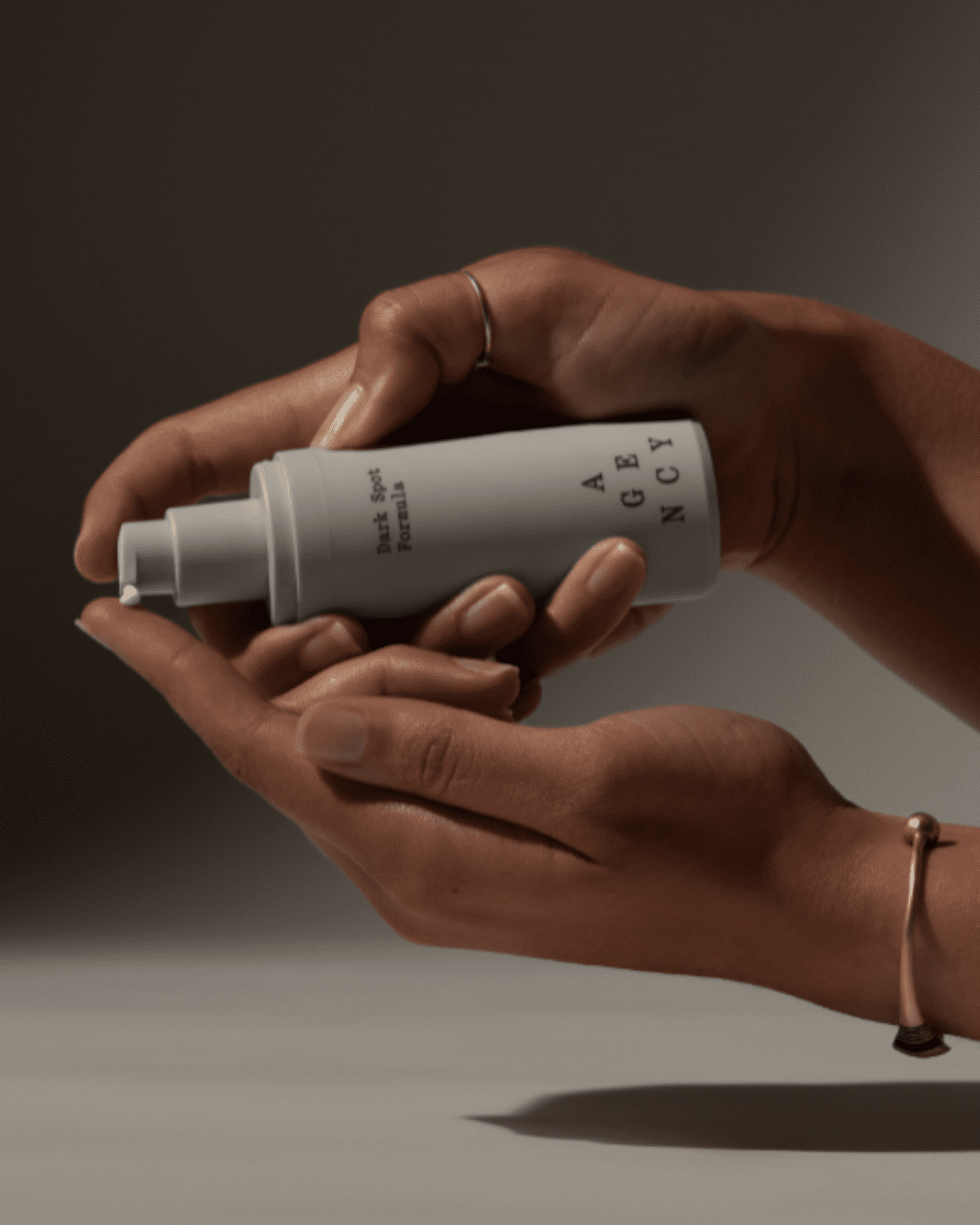 Two hands demonstrating the use of Agency Skincare Dark Spot Formula moisturizer; one hand presses the pump while the other hand's pointer finger collects the product, showing proper application.