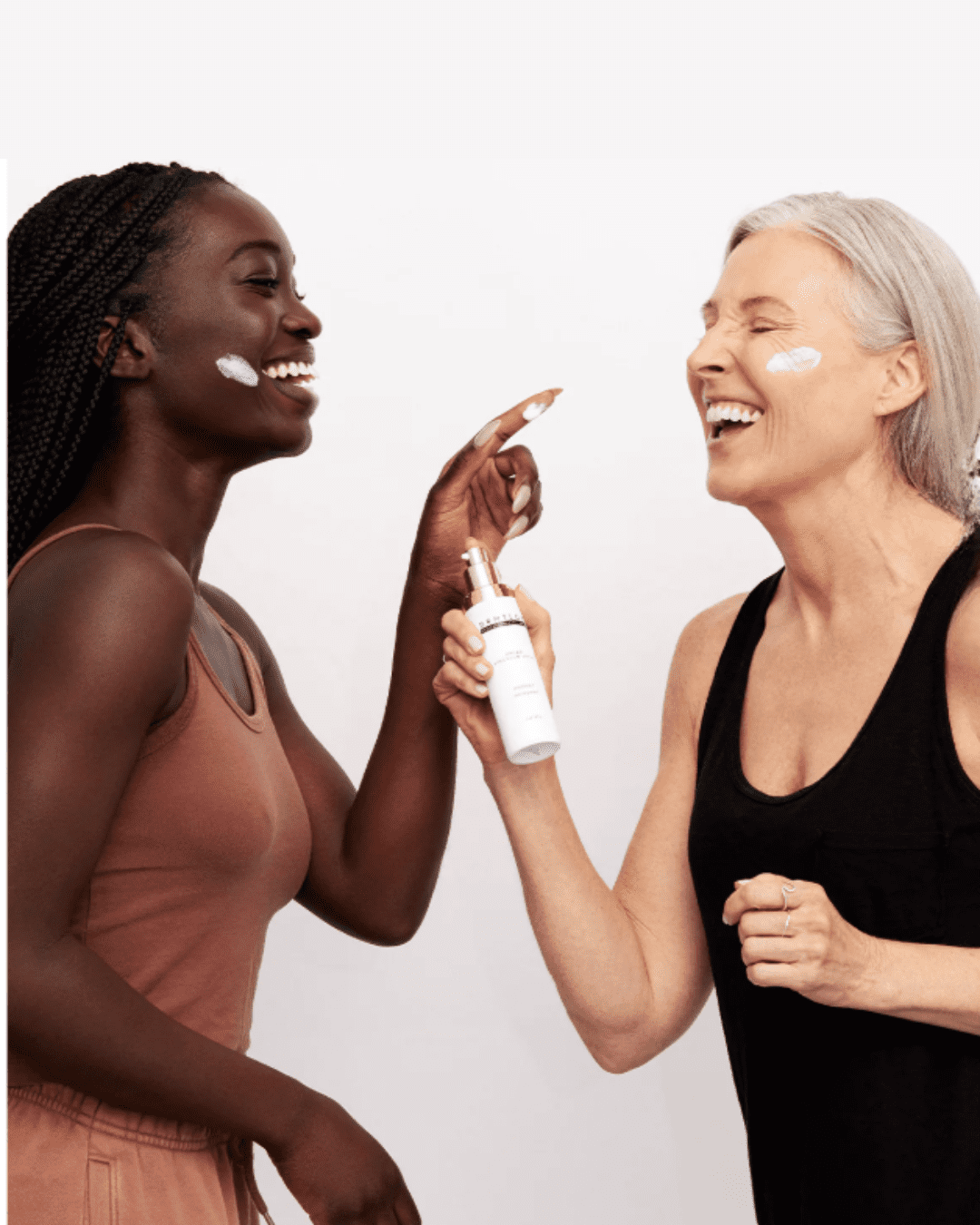 A young Black woman and an older white woman laughing together; the Black woman has a swatch of DRMTLGY Broad Spectrum SPF 45 on her face while the white woman holds the bottle, demonstrating sun protection and joyful skincare moments against a white background.