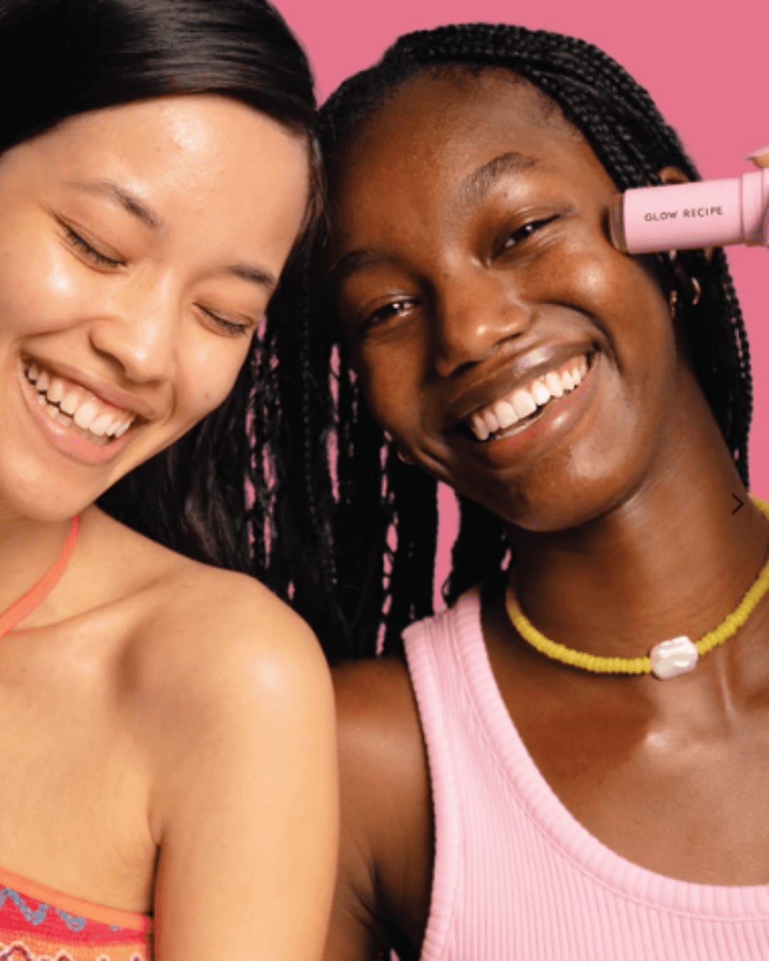 Two women, one Asian and one Black, holding Glow Recipe Watermelon Glow Niacinamide Dew Balm SPF 45 up to her face, as if applying the product, wearing colorful tops and jewelry, highlighting acne safe sunscreen use.