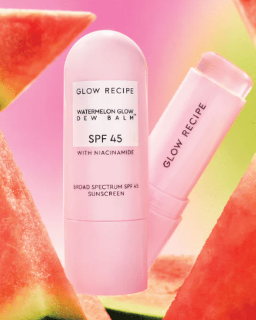 Glow Recipe Watermelon Glow Niacinamide Dew Balm SPF 45 standing upright next to an open tube showing the rolled-up product, surrounded by watermelon slices on a pink-to-yellow ombre background, demonstrating acne sun protection.