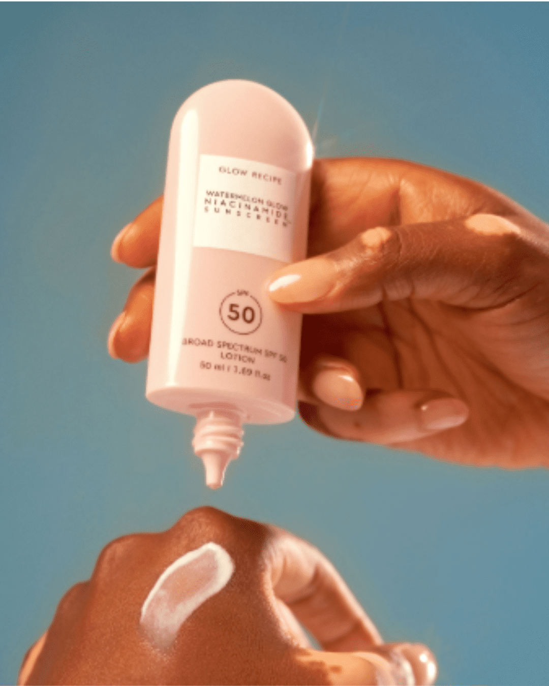 Black woman holding Glow Recipe Watermelon Glow Niacinamide Sunscreen SPF 50 with a swatch of the sunscreen on her hand, showing an acne safe sunscreen application on a blue-gray background.
