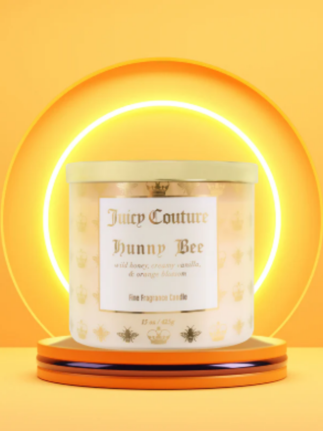 Juicy Couture Honey Bee candle with a gold lit displayed on a yellow platform against layered half-circle yellow backdrops with glowing light behind it.