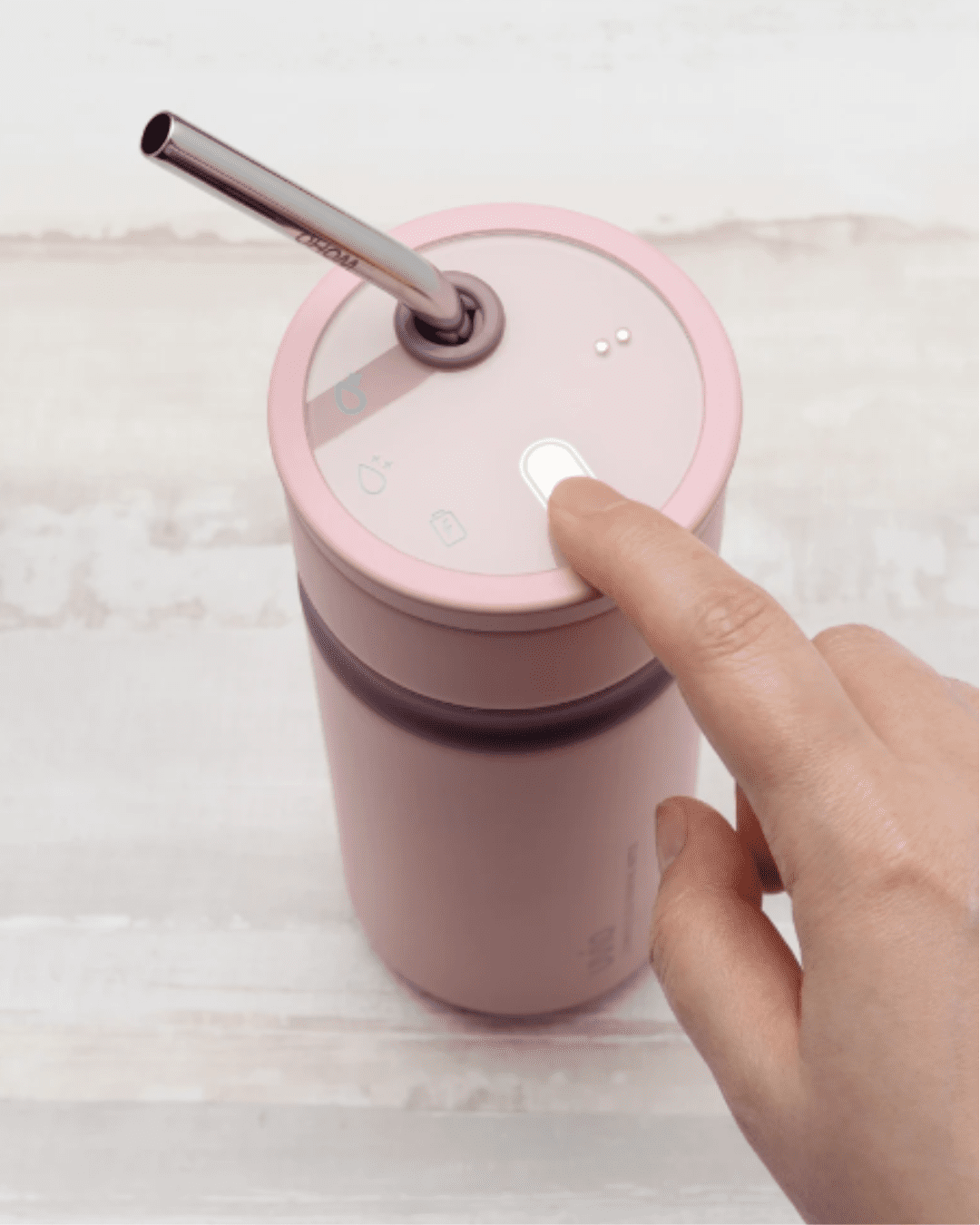 Overhead view of pink OHOM Self-Purifying Straw Water Bottle on a white marble surface, with a woman pressing the white lid to activate the purification system; a stainless-steel straw extends from the top.