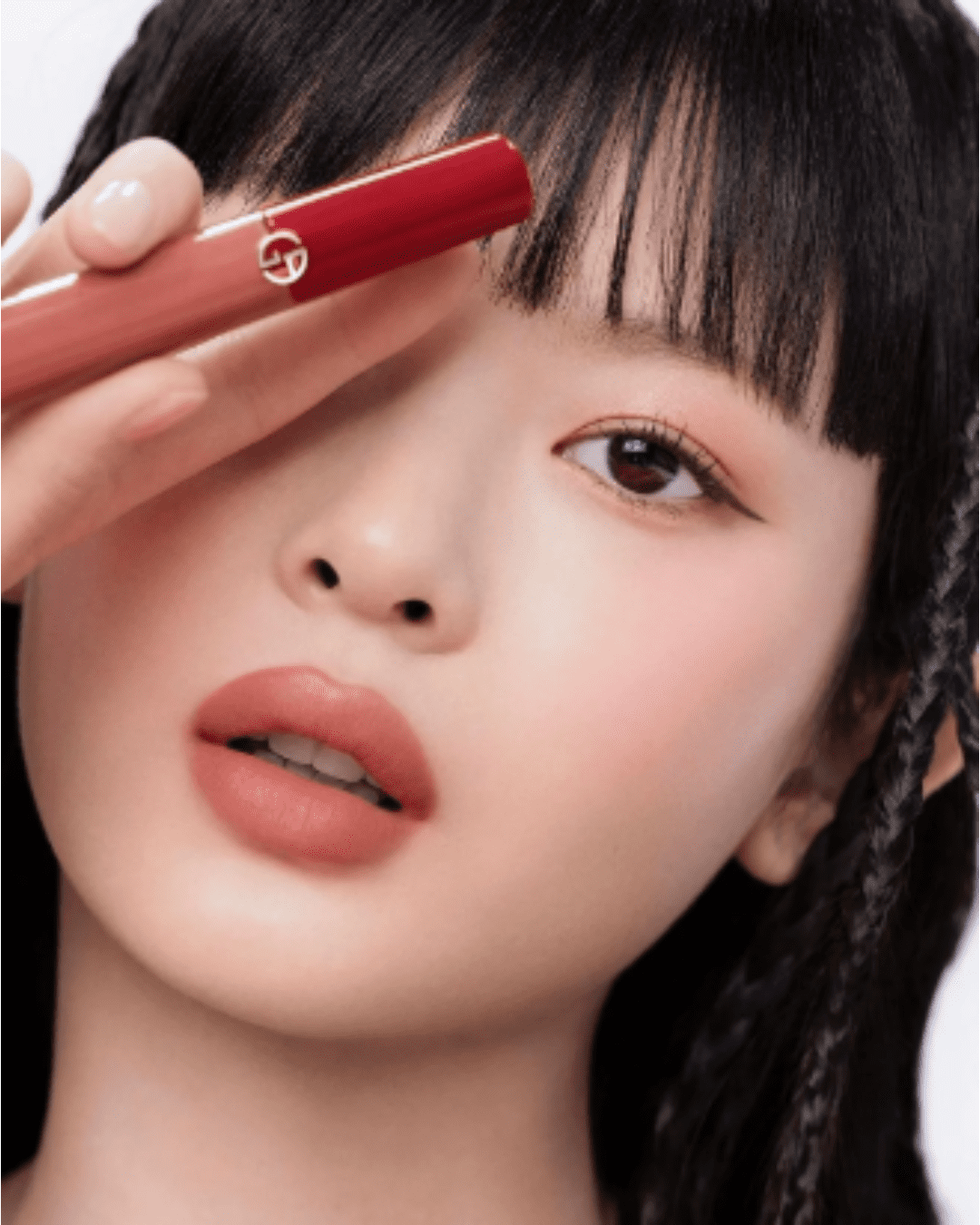 Hanni from NewJeans holding Giorgio Armani Lip Maestro Velvet Liquid Lipstick in shade 109 Tan up to her eye, showing packaging and color.