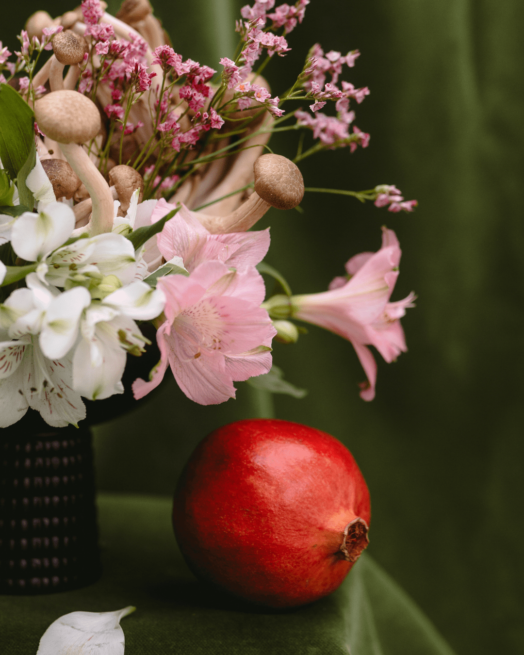 A textured green velvet tablecloth covers a small table topped with a wicker-style vase filled with a wild bouquet of pink and white flowers, mushrooms, and greenery, with a whole pomegranate placed beside it, styled as atmospheric decor for a candle lifestyle photo.